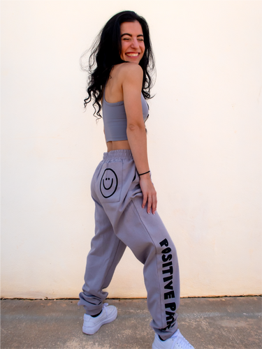 Peachy Pia Positive Pants joggers. Grey lightweight jogger sweatpants with positive pants design down the front of the right leg and smiley face design on the back pocket. Joggers provide an elastic waist and cuffed ankles.