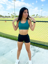 Load image into Gallery viewer, Young lady modeling Peachy Pia Worldwide Sports Bra. Sports bra is in color stone green with an 87% nylon, 13% spandex blend. Material is thick but flexible made to provide support and comfort.