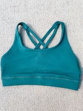 Load image into Gallery viewer, Young lady modeling Peachy Pia Worldwide Sports Bra. Sports bra is in color stone green with an 87% nylon, 13% spandex blend. Material is thick but flexible made to provide support and comfort.