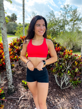 Load image into Gallery viewer, Young lady modeling Peachy Pia Worldwide Sports Bra. Sports bra is in color red with an 87% nylon, 13% spandex blend. Material is thick but flexible made to provide support and comfort.