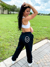 Load image into Gallery viewer, Young lady modeling Peachy Pia Positive Pants joggers. Black, fleece-lined joggers are made with a 100% cotton surface, coated in 100% polyester for increased durability with positive pants design down the front of the right leg and smiley face design on the back pocket. Joggers provide an elastic waist and cuffed ankles.