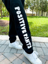 Load image into Gallery viewer, Young lady modeling Peachy Pia Positive Pants joggers. Black, fleece-lined joggers are made with a 100% cotton surface, coated in 100% polyester for increased durability with positive pants design down the front of the right leg and smiley face design on the back pocket. Joggers provide an elastic waist and cuffed ankles.