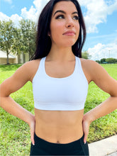 Load image into Gallery viewer, Young lady modeling Peachy Pia Worldwide Sports Bra. Sports bra is in color white with an 87% nylon, 13% spandex blend. Material is thick but flexible made to provide support and comfort.