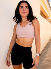 Load image into Gallery viewer, Young lady modeling a ribbed, beige sports bra that has medium/high support with a v neck front and scoop neck back.
