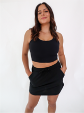 Load image into Gallery viewer, The Complimentary SweatSkirt has an outer skirt built with an elastic waistband and pockets. The Skirt has built-in spandex shorts and a happy message on the tag. Skirt is in color black.
