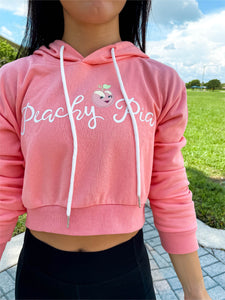 Young lady modeling Peachy Pia cropped hoodie. Cropped hoodie is Terry material with a 70% cotton, 30% polyester blend in a pinkish-peach color and has the official Peachy Pia logo on the front.