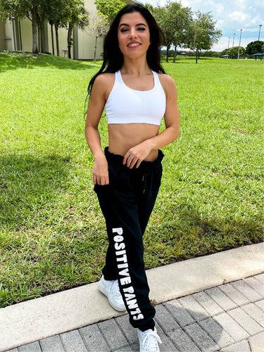 Young lady modeling Peachy Pia Positive Pants joggers. Black, fleece-lined joggers are made with a 100% cotton surface, coated in 100% polyester for increased durability with positive pants design down the front of the right leg and smiley face design on the back pocket. Joggers provide an elastic waist and cuffed ankles. 